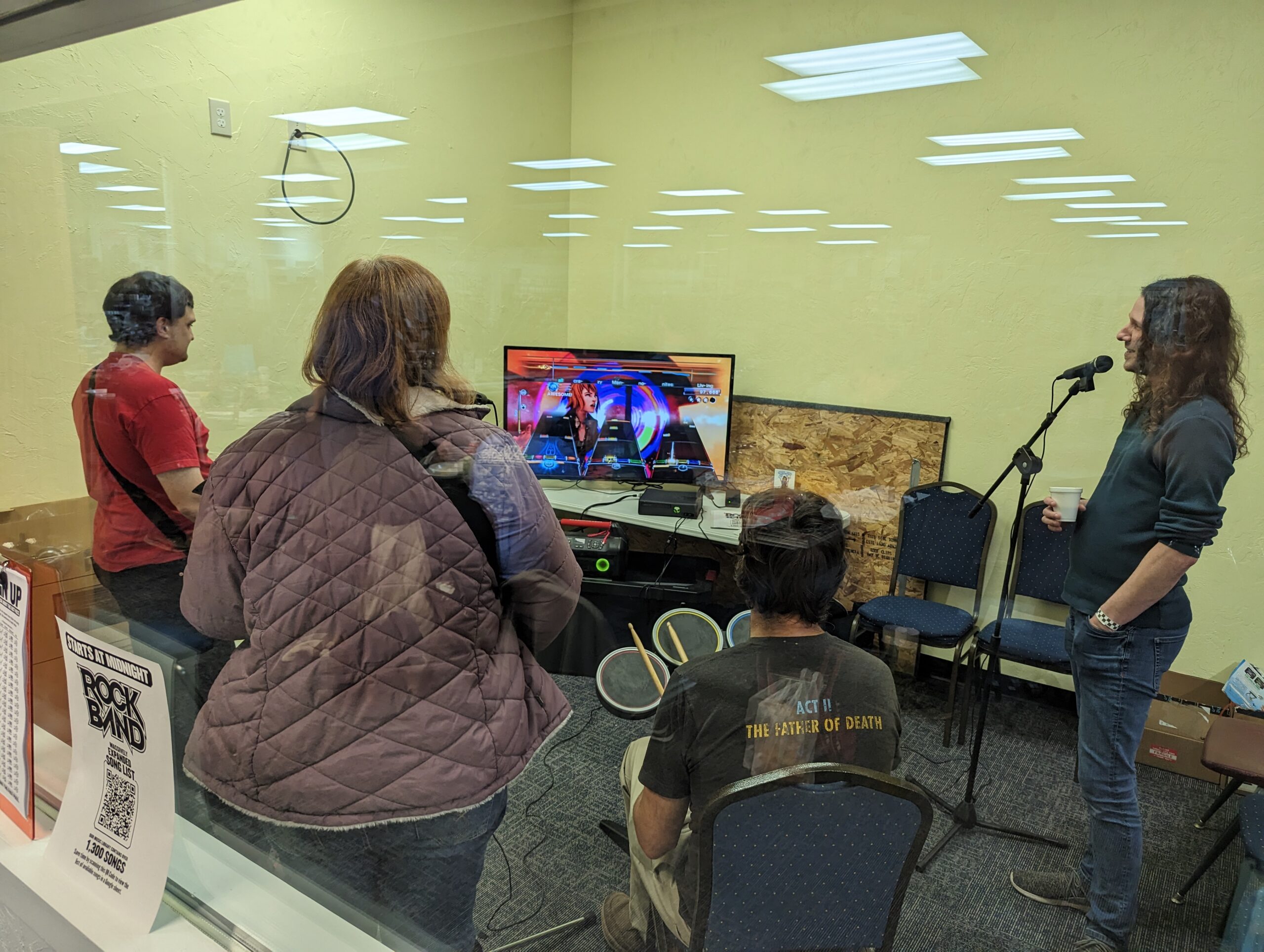 Four people play the game Rock Band with various instruments and a song on TV.