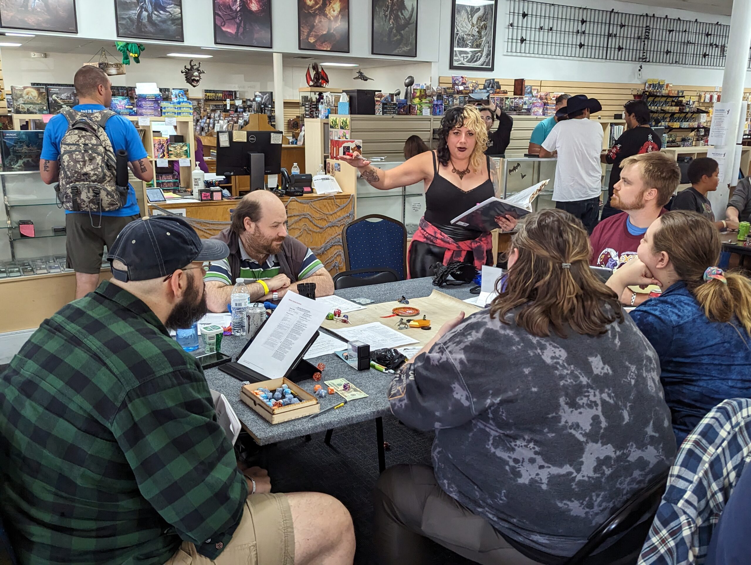 A woman stands while animatedly playing a game of D&D with a 5 other people around the table looking at her.