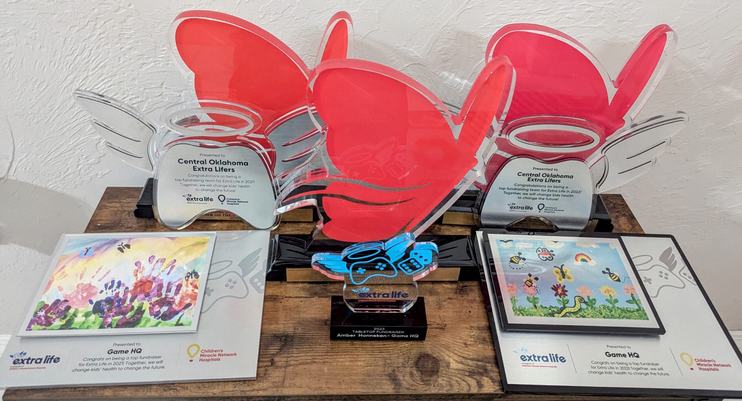 A collection of awards for Extra Life some have a butterfly motifs, others a game controller with wings and two feature children's artwork